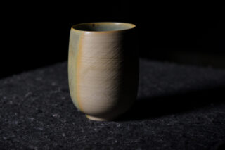 Eine einzelne Keramiktasse mit matter Oberfläche steht auf einer dunklen, strukturierten Oberfläche, wird von der Seite beleuchtet und hat einen Schatten auf der Rückseite.