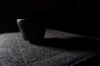 Eine kleine, dunkle Tasse steht auf einer strukturierten Oberfläche, wird von der Seite beleuchtet und wirft in einer schwach beleuchteten Umgebung einen Schatten.