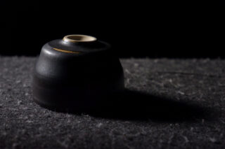 Eine schwarze Keramikschale steht verkehrt herum auf einer dunklen, strukturierten Oberfläche, wird von Licht von links beleuchtet und wirft einen Schatten nach rechts.