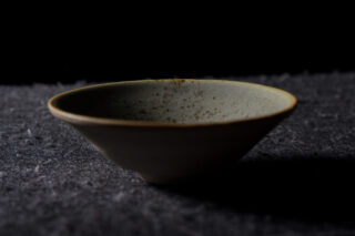 Eine flache, runde Keramikschale mit einer gesprenkelten hellen Glasur steht auf einer dunklen strukturierten Oberfläche vor einem schwarzen Hintergrund.