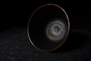 Eine dunkle Keramikschale mit metallisch schillernder Glasur auf der Innenseite liegt auf der Seite vor einem schwarzen Hintergrund.