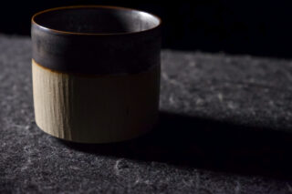 Eine Keramiktasse mit dunkler oberer und heller unterer Hälfte steht auf einer strukturierten, grauen Oberfläche und wirft einen Schatten nach rechts.