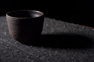 Eine einzelne dunkle Keramiktasse steht auf einer strukturierten Oberfläche und wirft bei schwachem Licht einen Schatten nach rechts.