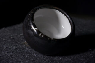 Eine kleine, runde, schwarze Keramikschüssel mit weißer Innenseite, die auf einer dunklen strukturierten Oberfläche auf die Seite geneigt ist.