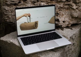 Auf einem Laptop auf einer Steinoberfläche wird eine Webseite mit Produktdesignbildern angezeigt, darunter eine aufgerollte braune Tasche und ein dreieckiges Objekt.