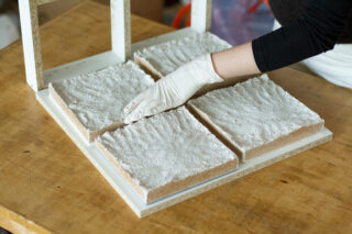 Eine Person mit einem weißen Handschuh ordnet vier rechteckige Platten, die mit einer weißen pulverförmigen Substanz beschichtet sind, auf einem Holztisch an.