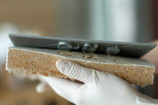 Eine behandschuhte Hand hält ein raues, rechteckiges Stück Isoliermaterial, gegen dessen Oberfläche ein Metallwerkzeug gedrückt wird.