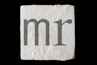 Eine weiße, strukturierte quadratische Fliese mit den in Grau aufgedruckten Kleinbuchstaben „mr“ auf der Oberfläche vor einem schwarzen Hintergrund.