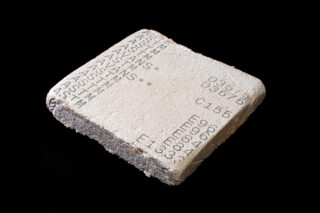 Ein quadratischer Betonblock mit eingravierten Buchstaben, Zahlen und Symbolen auf der Oberfläche, dargestellt vor einem schwarzen Hintergrund.