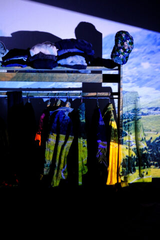 An einem Ständer hängen Kleidungsstücke, auf denen gefaltete Gegenstände liegen. Auf die Kleidungsstücke und die Wand wird eine Landschaft projiziert, die mit den Mustern der Kleidung verschmilzt.