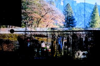 Eine Küchenarbeitsplatte mit Utensilien und Gläsern ist schwach beleuchtet, während im Hintergrund eine Landschaft aus Bäumen und Bergen projiziert wird.