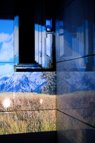 Ein Bild einer Wand und eines offenen Fensters mit einer projizierten Szene aus Bergen und einem Feld auf den Oberflächen, wodurch im Innenbereich die Illusion einer Landschaft im Freien entsteht.