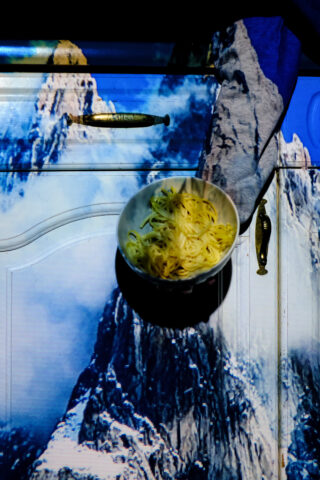 Auf einer Schranktür steht eine Schüssel Pasta. Auf die Tür und die Pasta wird eine Bergszene projiziert, wodurch sich Texturen und Farben vermischen.