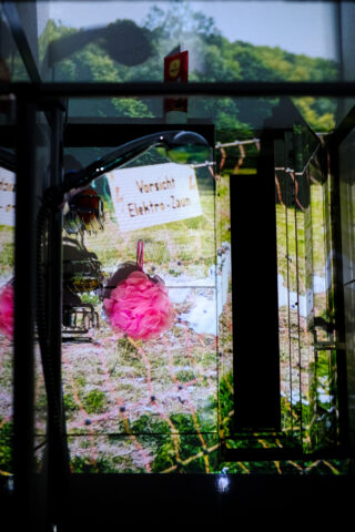 Ein Foto einer Duschkabine mit einem Luffaschwamm, auf das ein Bild eines Warnschildes „Vorsicht Elektro-Zaun“ und eine Landschaftsszene projiziert wird.