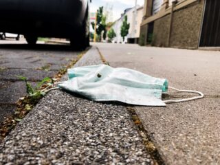 Eine Einweg-Gesichtsmaske liegt weggeworfen auf einem Bürgersteig neben dem Bordstein, im Hintergrund sind ein Auto und etwas Grün zu sehen.