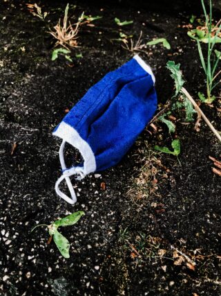 Eine blaue Gesichtsmaske liegt weggeworfen auf dunklem, feuchtem Boden mit verstreuten grünen Pflanzen und trockenem Gras.