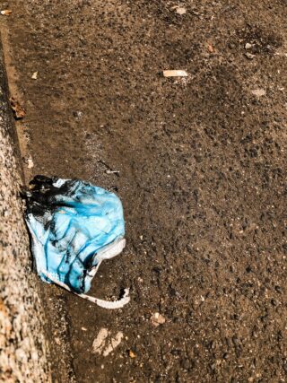 Eine weggeworfene blaue Gesichtsmaske liegt auf einem rauen, schmutzigen Bürgersteig neben einem Bordstein und ist teilweise mit einer dunklen Substanz befleckt.