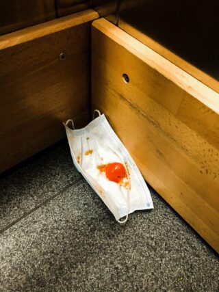 Eine Einweg-Gesichtsmaske mit einer zerbrochenen Kirschtomate und Soße darauf liegt in der Ecke auf dem Boden vor Holzpaneelen.