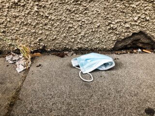 Eine blaue Einweg-Gesichtsmaske liegt zusammen mit etwas getrocknetem Gras und Schutt auf einem Betongehweg in der Nähe einer strukturierten Wand.