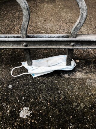 Eine Einweg-Gesichtsmaske steckt unter einem Metallzaun auf einer rauen Betonoberfläche fest.