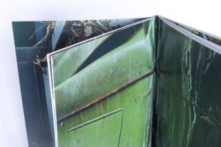 Auf einer weißen Fläche liegt ein offenes Buch, das Nahaufnahmen einer grünen, verrosteten Metalloberfläche und von Trümmern zeigt.