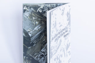 Ein teilweise geöffnetes Buch, das eine Seite mit abstrakten metallischen Texturen und eine andere mit weißen und grauen Mustern und deutschem Text zeigt.