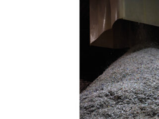 Eine große Industrierutsche lädt einen Haufen zerkleinerter Materialien, möglicherweise Glas oder Kies, auf einem Hügel in einer schwach beleuchteten Anlage ab.