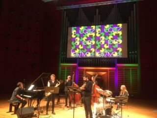 Auf der Bühne tritt eine Jazzband mit Klavier, Saxophon, Trompete, Kontrabass, Schlagzeug und Gitarre auf, über ihnen prangt ein buntes „Jazz Festival“-Schild.