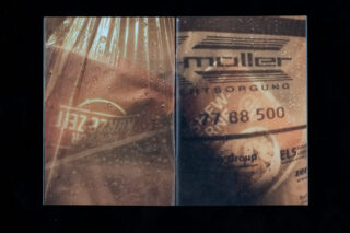 Eine Nahaufnahme von zwei teilweise verdeckten Schildern hinter einer regennassen Oberfläche, mit deutschem Text und einer Telefonnummer auf der rechten Seite.