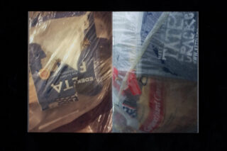 Vor dunklem Hintergrund stehen zwei in Plastikfolie eingewickelte Packungen nebeneinander, eine mit der Aufschrift „EDEKA Feta“ und die andere mit der Aufschrift „Cracker“ und Brot.