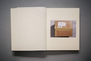 In einem offenen Buch ist auf der rechten Seite zentriert das Foto eines kleinen Kartons mit einem Versandetikett zu sehen.