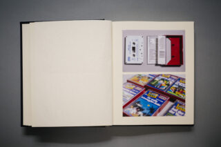 In einem offenen Buch ist das Foto einer Kassette mit Verpackung über einem Bild mehrerer diagonal angeordneter bunter VHS-Kassettenschachteln zu sehen.