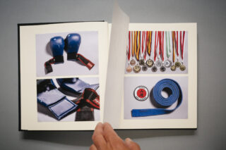 Eine Hand blättert durch ein Sammelalbum mit Fotos von blauen Kampfsporthandschuhen, Fußschützern, einem blauen Gürtel, einem Aufnäher und einer Sammlung von Medaillen mit bunten Bändern.