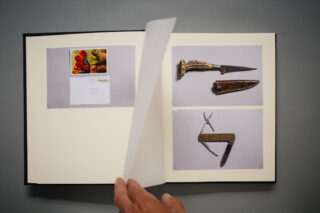 Ein aufgeschlagenes Buch zeigt Fotos eines Umschlags mit einer Karte, eines dekorativen Dolches mit Scheide und eines mehrklingen Taschenmessers. Eine Hand blättert darin.