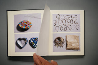 Eine Hand blättert in einem Fotobuch, das Bilder eines bunten runden Ornaments, herzförmiger Gegenstände, Metallhaken, einer aufgerollten Schnur und eines verpackten Pakets auf grauem Hintergrund zeigt.