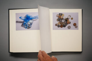 Ein offenes Buch mit zwei Fotos: eines von einer blauen Plastiktüte mit Inhalt und ein anderes von verschiedenen Münzen, wobei eine Hand die Seite umblättert.