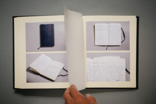 Eine Hand blättert in einem offenen Fotoalbum, das Bilder eines schwarzen Notizbuchs und handschriftliche Notizen auf grauem Hintergrund zeigt.