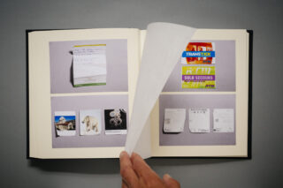Eine Hand blättert eine Seite in einem Fotobuch um, das Bilder von Papiergegenständen wie Notizen, Tickets, Karten und Quittungen auf einem grauen Hintergrund zeigt.