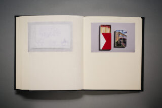 Ein offenes Buch, das auf der linken Seite ein durchscheinendes Blatt und auf der rechten Seite ein Foto einer Streichholzschachtel mit einem Gebäudebild zeigt.