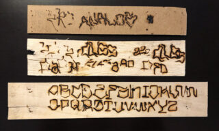Auf drei Holzbrettern sind eingebrannte Inschriften zu sehen; oben sind Symbole und Buchstaben, in der Mitte abstrakte Zeichen und unten ein stilisiertes Großbuchstabenalphabet zu sehen.