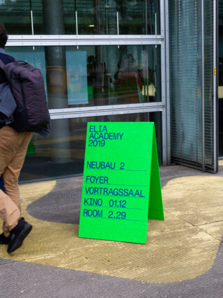 Ein leuchtend grünes Schild zeigt die Veranstaltungshinweise zur ELIA Academy 2019 und listet Standorte wie Neubau 2, Foyer, Vortragssaal, Kino 01.12 und Raum 2.29 in der Nähe eines Gebäudeeingangs auf.