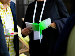 Zwei Personen stehen bei einer Veranstaltung zusammen. Eine hält eine Broschüre mit grünen Seiten, die andere einen Teller mit einer Tasse und bunten Süßigkeiten. Beide tragen Namensschilder.