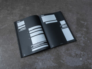 Auf einer strukturierten Oberfläche liegt ein offenes Buch, das zwei schwarze Seiten mit verschiedenen weißen rechteckigen Formen und minimalen Textelementen zeigt.