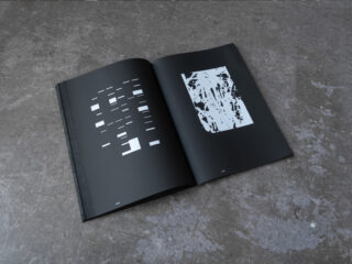 Ein offenes schwarzes Buch zeigt auf der linken Seite abstrakte weiße rechteckige Formen und auf der rechten Seite ein komplexes weißes Muster, das auf einer strukturierten grauen Oberfläche liegt.