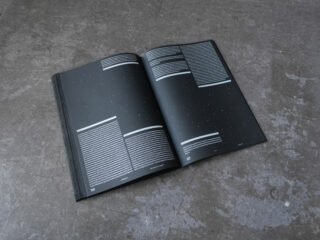 Eine aufgeschlagene Zeitschrift mit schwarzen Seiten, weißen Textblöcken und kleinen weißen Punkten, auf einer Betonoberfläche liegend.