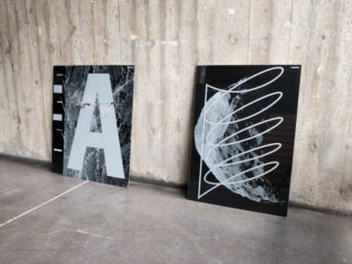 Zwei gerahmte abstrakte Drucke lehnen an einer strukturierten Betonwand; einer zeigt einen großen Buchstaben „A“, der andere ein wirbelndes Linienmuster.