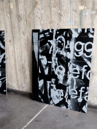 Eine große rechteckige Tafel mit abstrakten schwarz-weißen Mustern und teilweise sichtbaren Buchstabenfragmenten, die an einer verwitterten Holzwand lehnt.