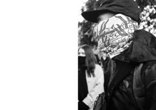 Eine Person mit Mütze und gemusterter Mund-Nasen-Bedeckung steht im Freien inmitten einer Gruppe von Menschen, von denen einige ebenfalls Masken tragen. Die linke Bildseite ist leer.