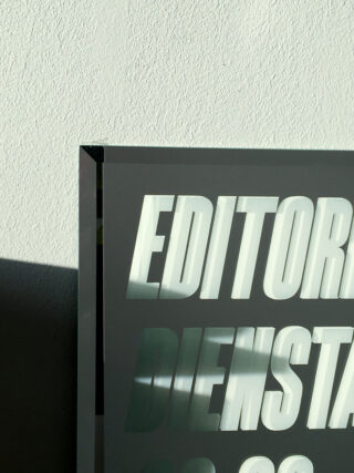 Eine Teilansicht eines Glasschildes mit großem weißem Text, einschließlich der sichtbaren Wörter „EDITOR“ und „DIENST“, vor einer strukturierten Lichtwand mit Schatten.
