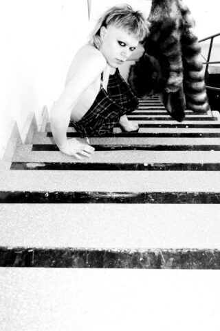 Eine Person mit kurzen Haaren und dramatischem Augen-Make-up sitzt barfuß auf einer Treppe. Sie trägt ein kariertes Outfit und einen Pelzmantel, der über das Geländer hängt. Schwarz-Weiß-Foto.